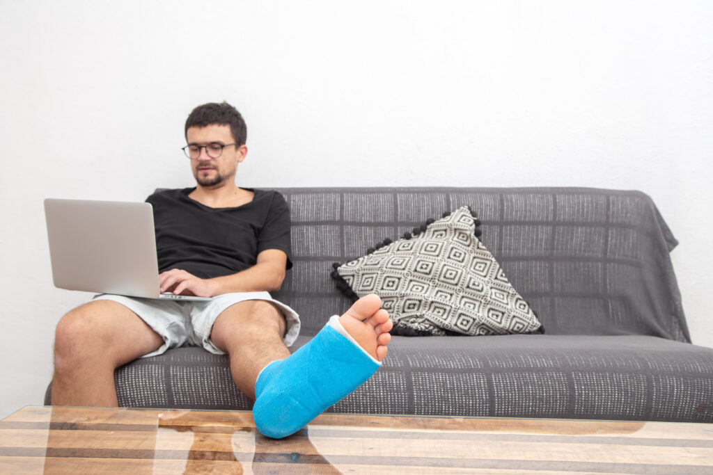 Homme avec une jambe dans le plâtre travaillant sur son ordinateur portable depuis son canapé