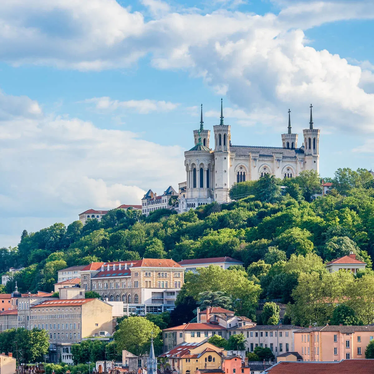 La basilique Notre-Dame de Fourvière, un imposant édifice blanc, surplombe la ville de Lyon verdoyante sous un ciel bleu parsemé de nuages blancs.